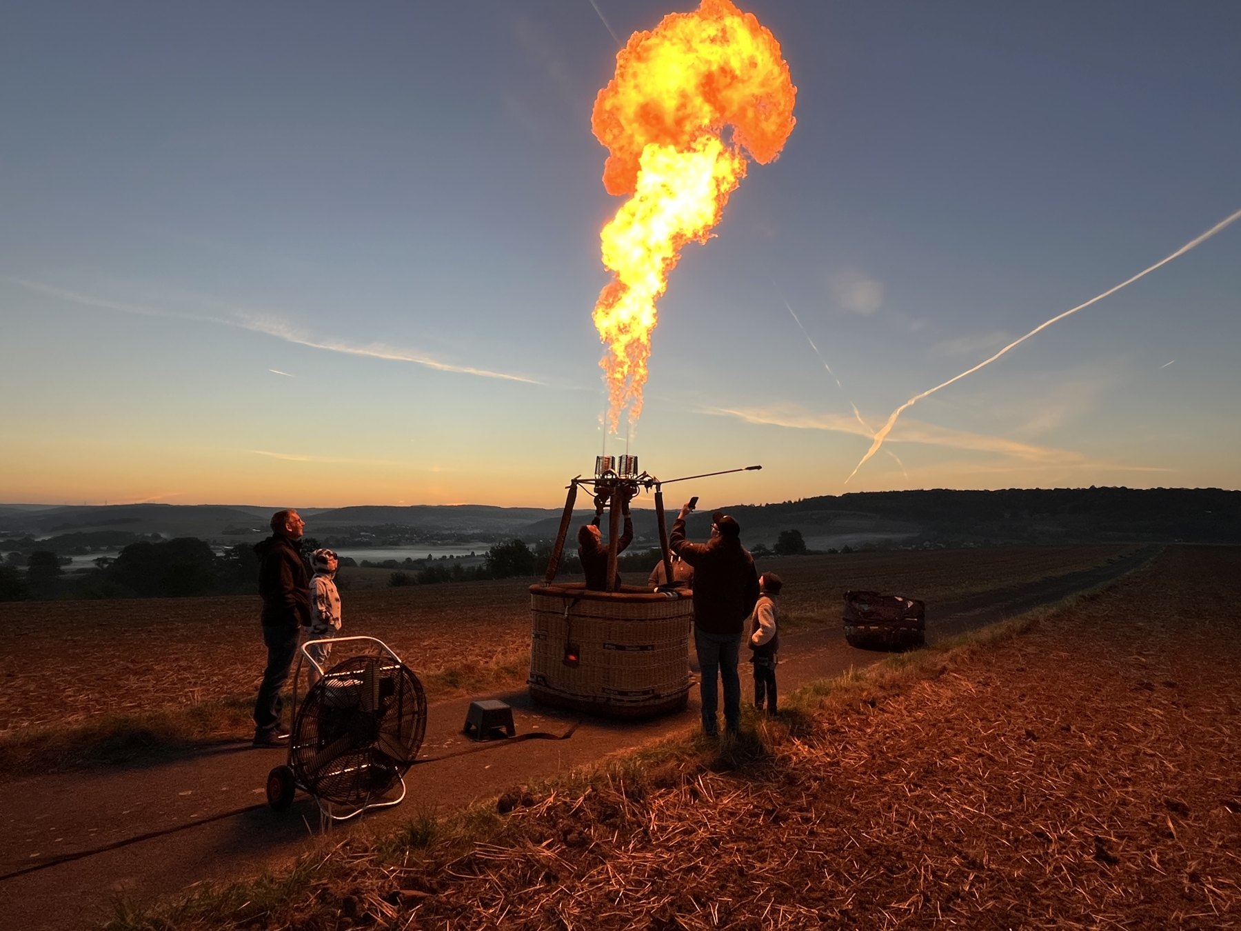 Mehrere Menschen am Ballonkorb mit leuchtendem Brenner vor der Landschaft im Abendlicht